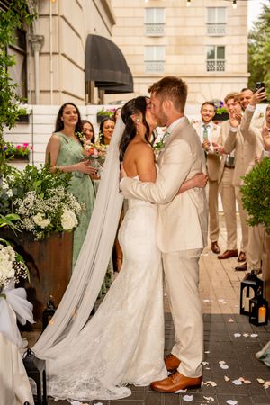 Bride and groom share first kiss at outdoor wedding ceremony with bridal party and guests celebrating behind them.