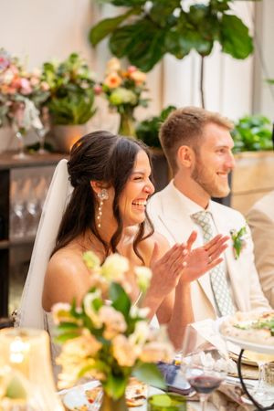 Bride laughing and clapping at wedding reception table with groom in beige suit, surrounded by floral arrangements.