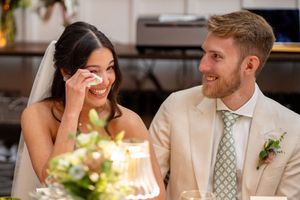 Bride wiping tears of joy while smiling beside groom in tan suit at wedding reception table.