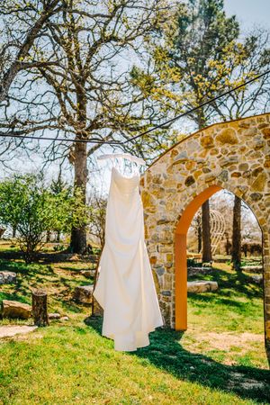 White wedding dress hanging outdoors near a stone arch and trees on a sunny day.