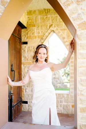 Bride in white strapless gown poses in stone arch doorway, wearing tiara and pearl jewelry on wedding day.