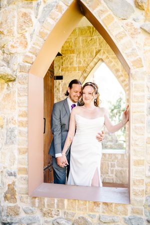 Bride and groom pose in a Gothic stone arch doorway, holding hands in wedding attire at a rustic venue.
