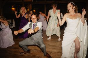 Bride and groom dance energetically with wedding guests on a wooden dance floor during a lively reception celebration.