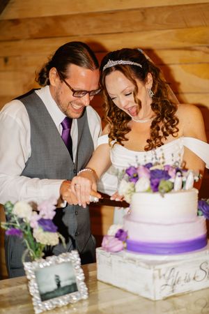 Bride and groom cutting a three-tier white and purple wedding cake together, smiling joyfully in a rustic wood-paneled venue.