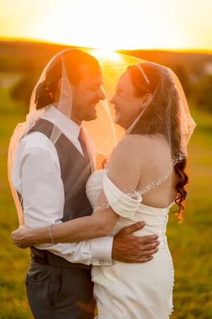 Bride and groom embrace under veil at golden sunset, smiling joyfully in outdoor wedding portrait.