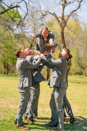 Groomsmen in gray suits tossing the groom into the air outdoors on a sunny day.