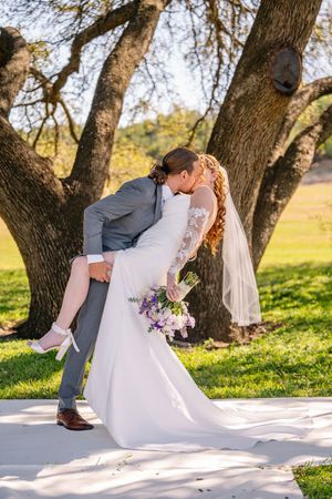 Groom dips bride for a romantic kiss outdoors under a large oak tree, bride holding purple floral bouquet in white gown.