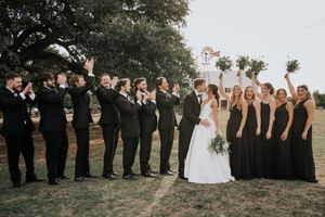 Wedding party in formal black attire poses outdoors for group photos at sunset.