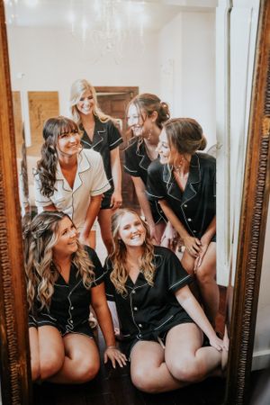 Bridesmaids in matching black pajama sets laughing together in mirror reflection.