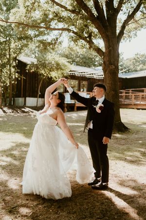 A bride and groom dance playfully under large trees during their outdoor wedding photoshoot.