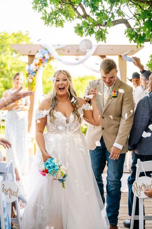 Joyful bride and groom walk down the aisle after wedding ceremony, bride holding colorful bouquet under floral arch.