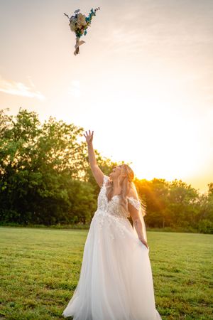 Bride in white lace gown tossing bouquet at sunset in green field.