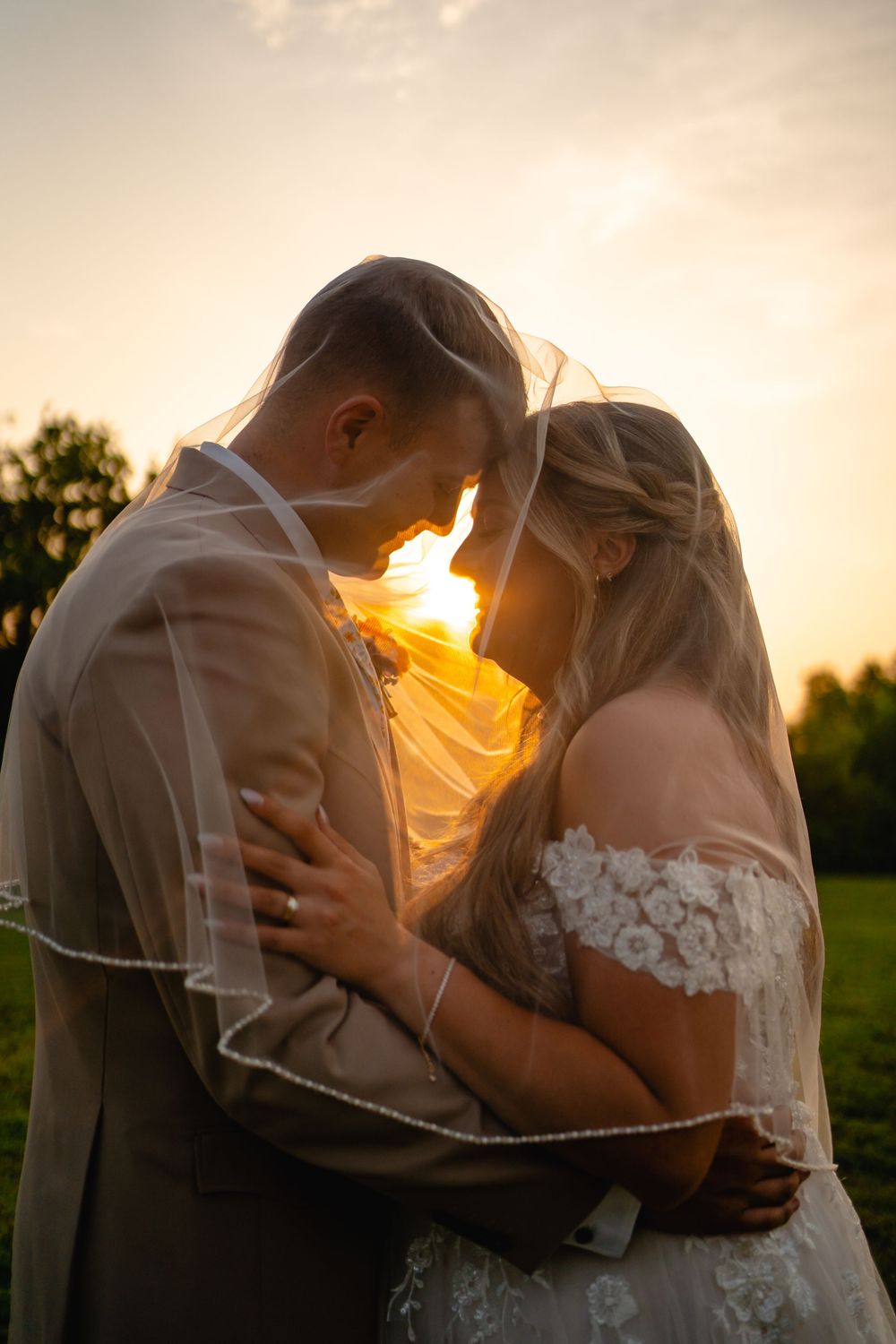 Bride and groom embrace under veil at golden sunset in lush green field, romantic wedding portrait.
