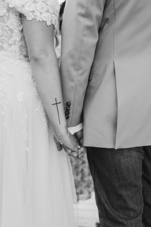 Black and white photo of bride and groom holding hands, bride has a cross tattoo on her forearm, wearing wedding attire.