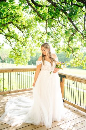 Bride in off-shoulder white tulle gown holding colorful bouquet on wooden deck under lush green tree canopy.
