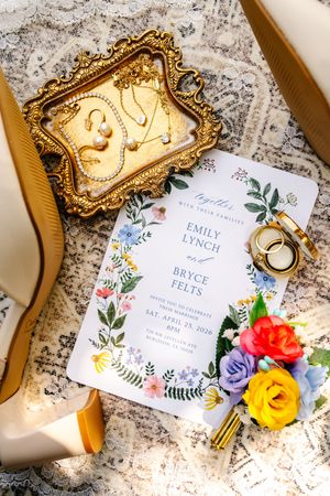 Wedding flat lay featuring floral invitation for Emily Lynch and Bryce Felts, gold jewelry tray, rings, colorful boutonniere, and heels.