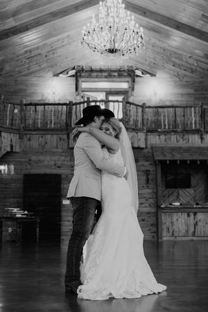 Black and white photo of a bride and groom sharing their first dance in a rustic barn venue with a chandelier overhead.