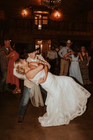 Groom dips bride in white lace gown during first dance at rustic barn wedding reception with guests watching.
