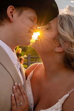 Bride and groom sharing a romantic sunset kiss, golden sunburst glowing between them, bride wearing lace gown and diamond ring.