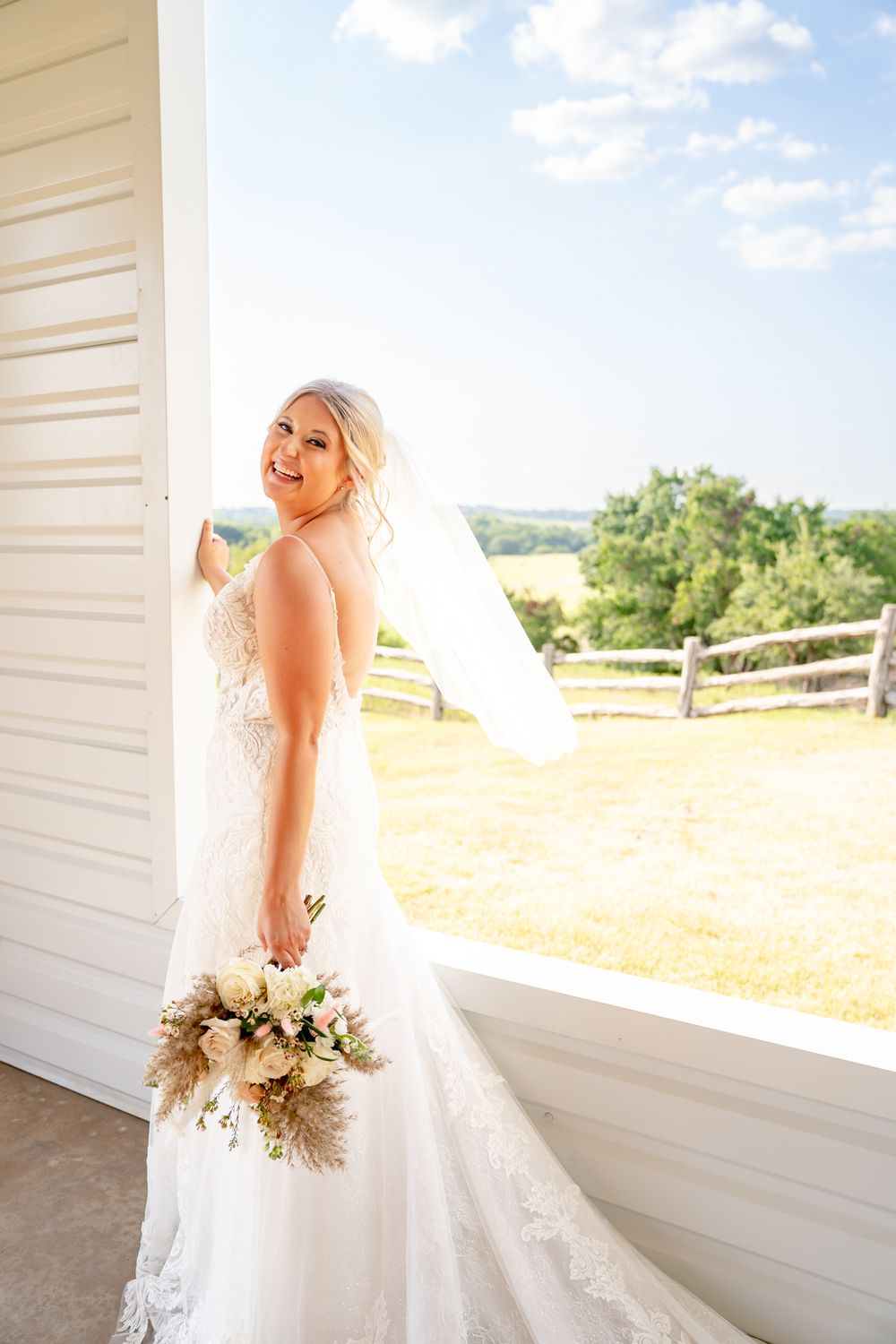 Smiling bride in lace gown holds bouquet by barn door, veil flowing, with sunny countryside backdrop.