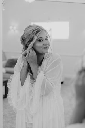 A bride in a white sheer robe adjusts her earring while smiling in a mirror during wedding preparations.