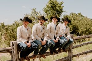 Four groomsmen in black cowboy hats and tan blazers sit on a rustic wooden fence outdoors.