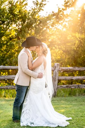 Bride and groom sharing a romantic kiss at golden hour, with rustic fence and glowing sunset backdrop.