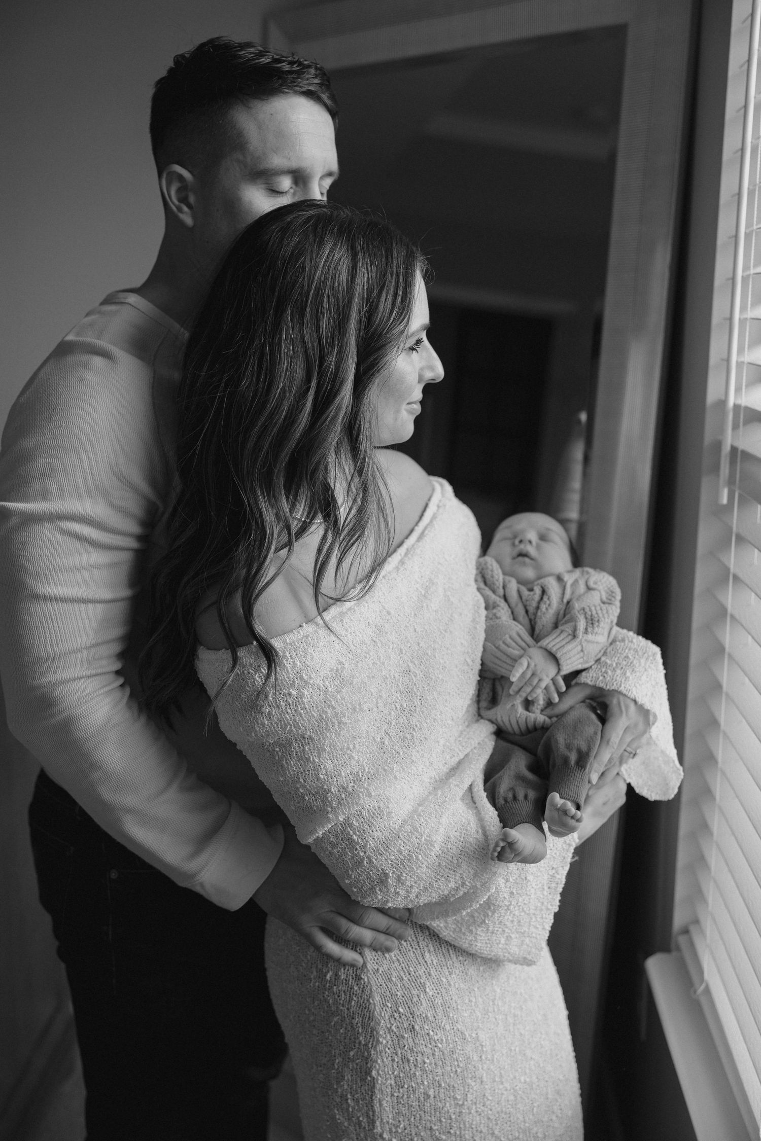 Family standing by window with newborn baby in cozy black and white portrait.