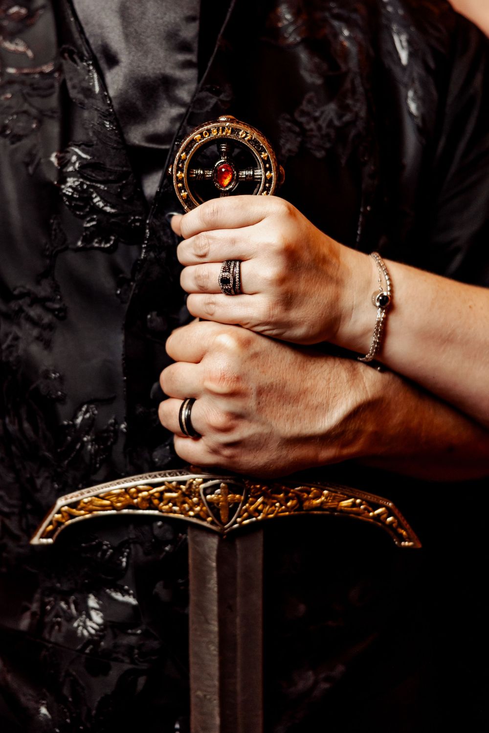 Close-up of hands wearing wedding rings holding an ornate compass on a decorative sword hilt.