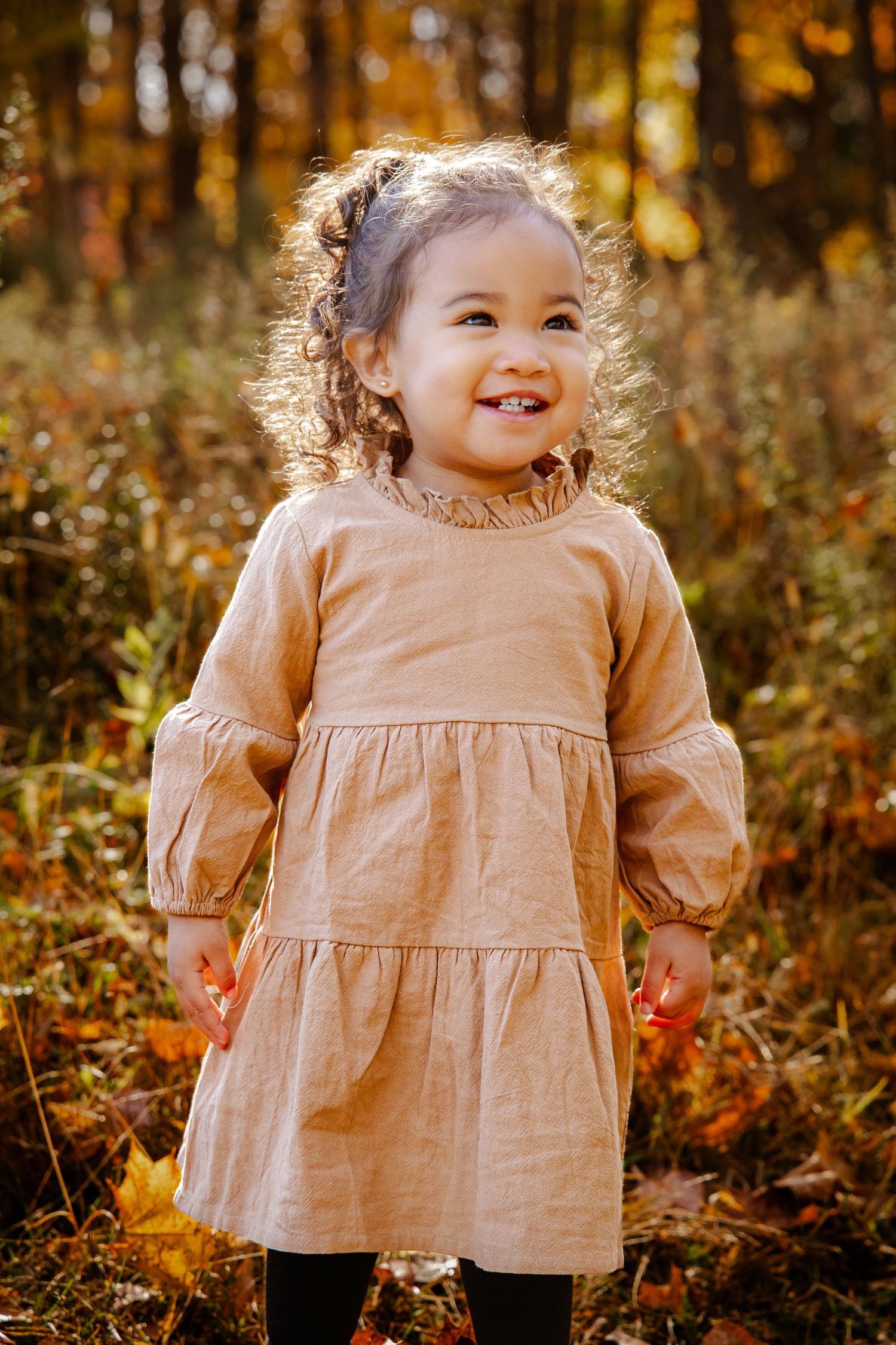 Small child in a beige dress playing among fallen autumn leaves.