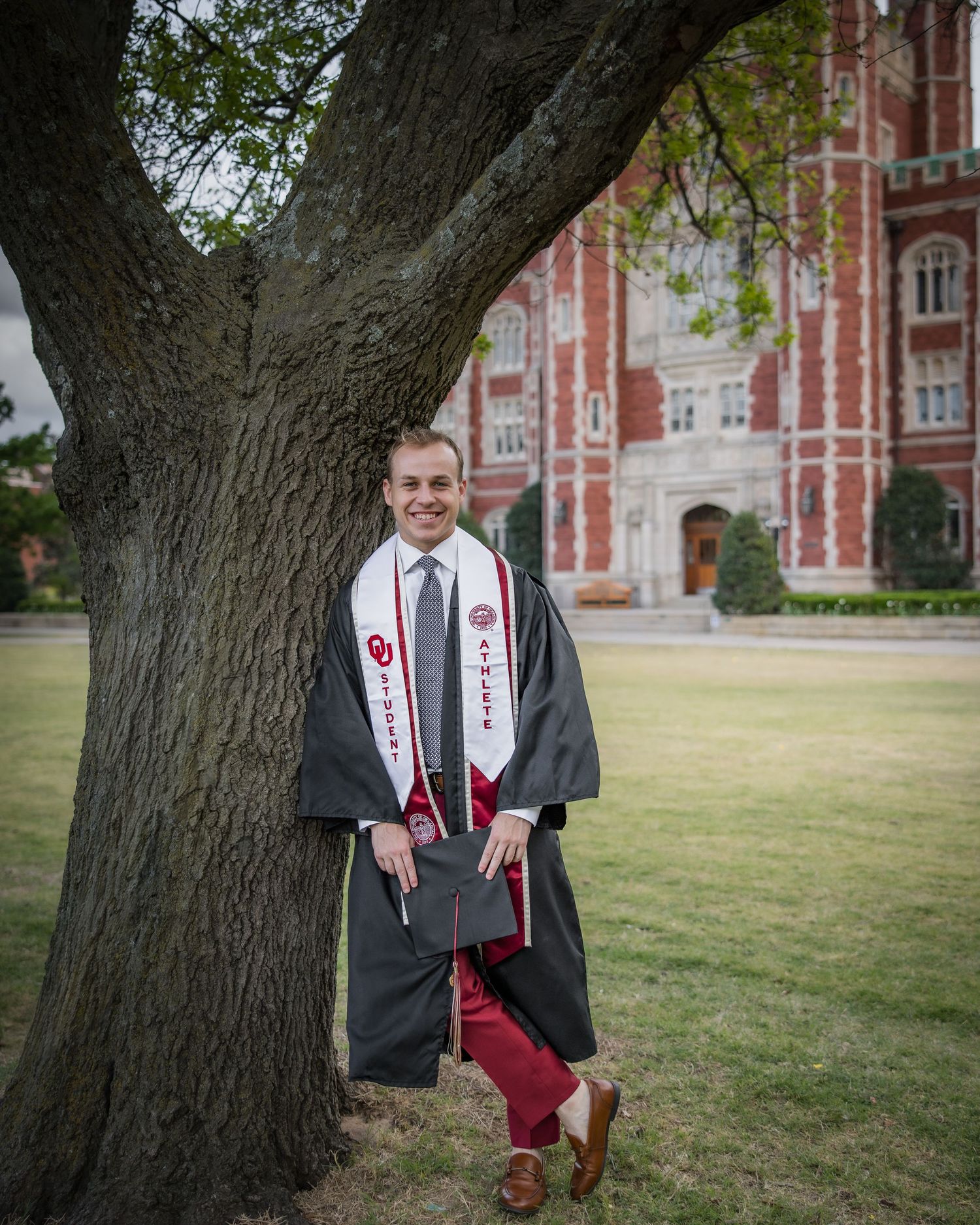 College graduate in cap and gown leans against tree outside red brick university building holding diploma.
