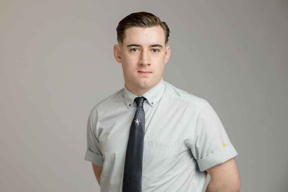 Professional portrait of person wearing a light gray short-sleeve button-down shirt with navy tie against neutral background.