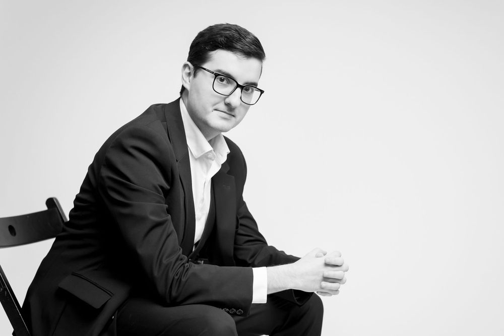 Black and white portrait of a professional in a suit and glasses sitting in a chair against a white background.