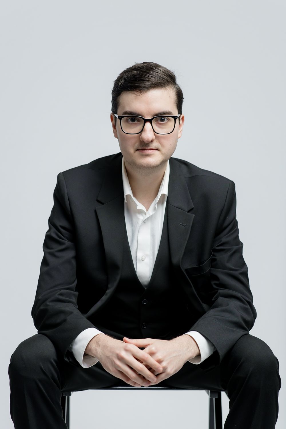 Professional studio portrait of a person in a dark suit and glasses seated on a white chair against a light background.