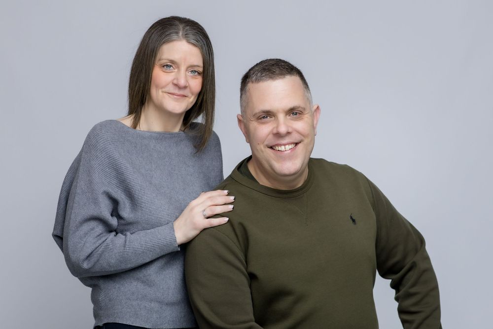 A couple wearing cozy sweaters pose together against a gray background, smiling warmly at the camera.