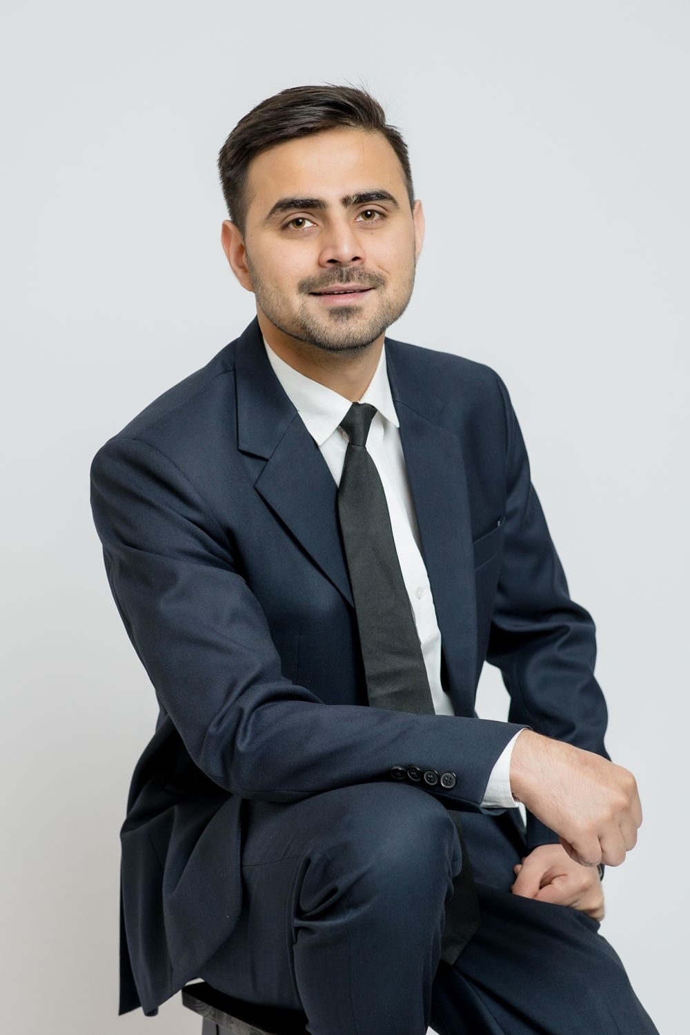 Professional portrait of someone in a dark suit and white tie sitting casually against light background.
