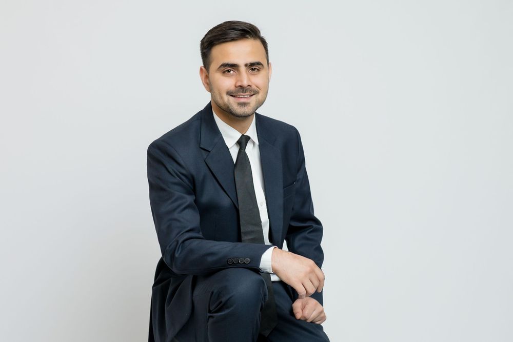 Professional in a dark navy business suit and tie sitting against a light gray background with a warm expression.