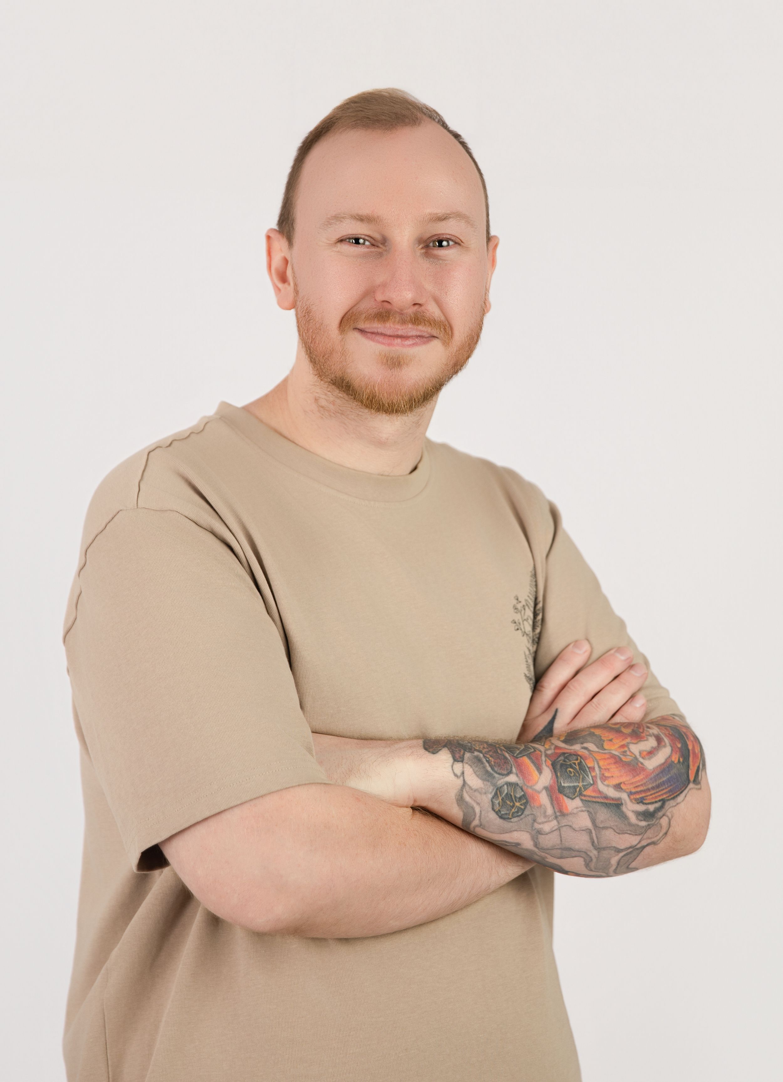 Professional portrait of someone in a beige t-shirt with visible arm tattoos standing with arms crossed against light background.