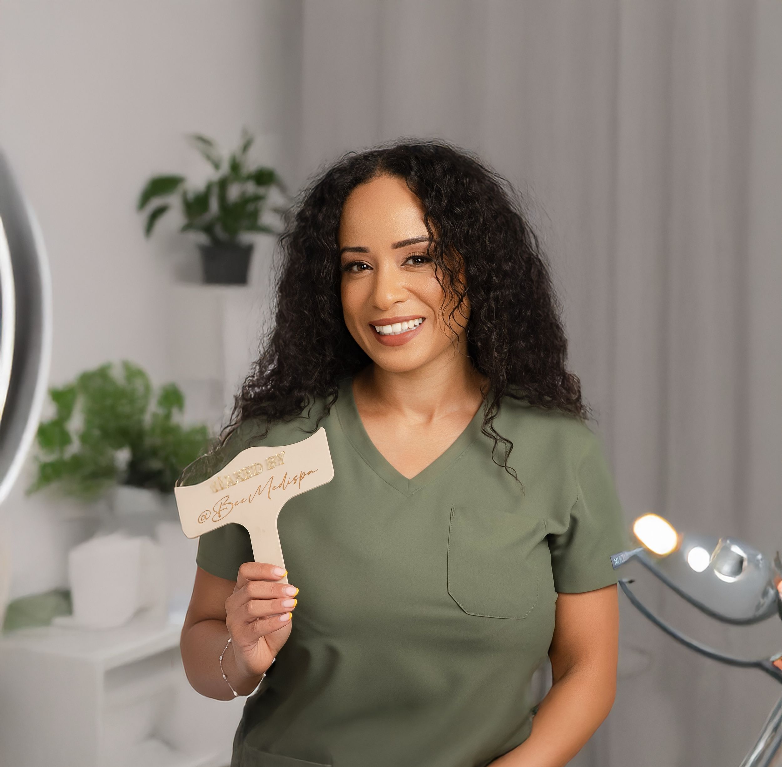 Person in olive green scrubs holds up a business card while setting up lighting and recording equipment in home studio.