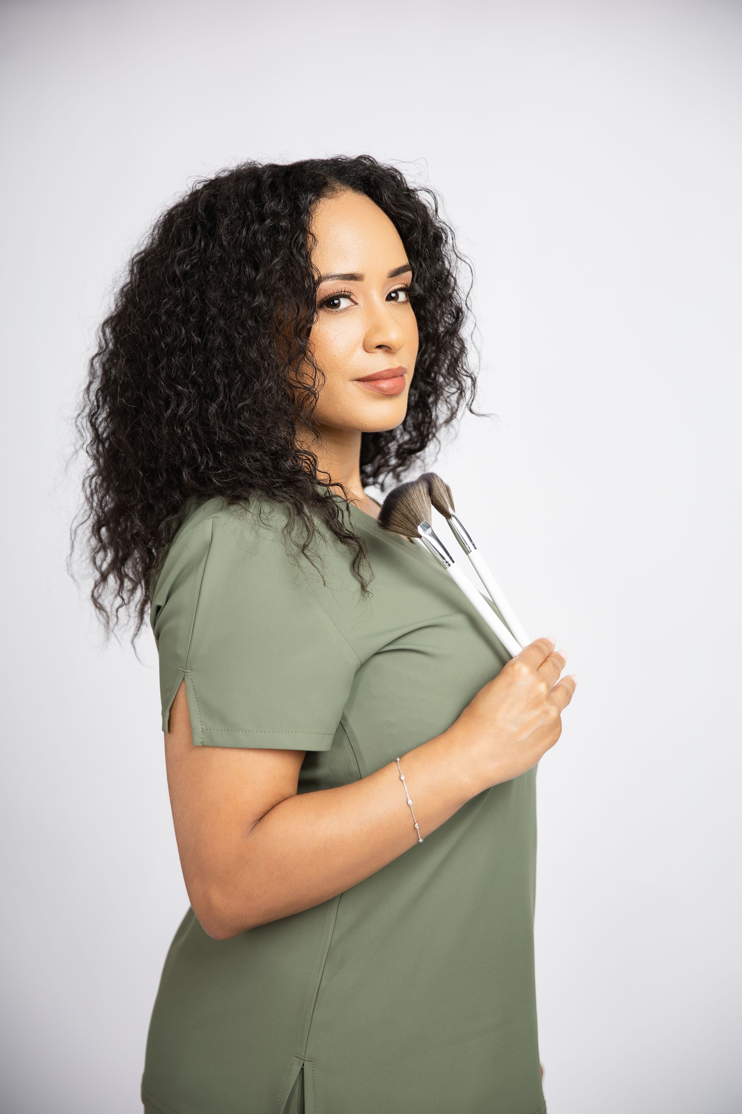 Professional with curly dark hair wearing sage green scrubs holds a makeup brush against light gray background.