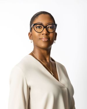 Professional headshot of person wearing black glasses and cream colored v-neck top against white background.