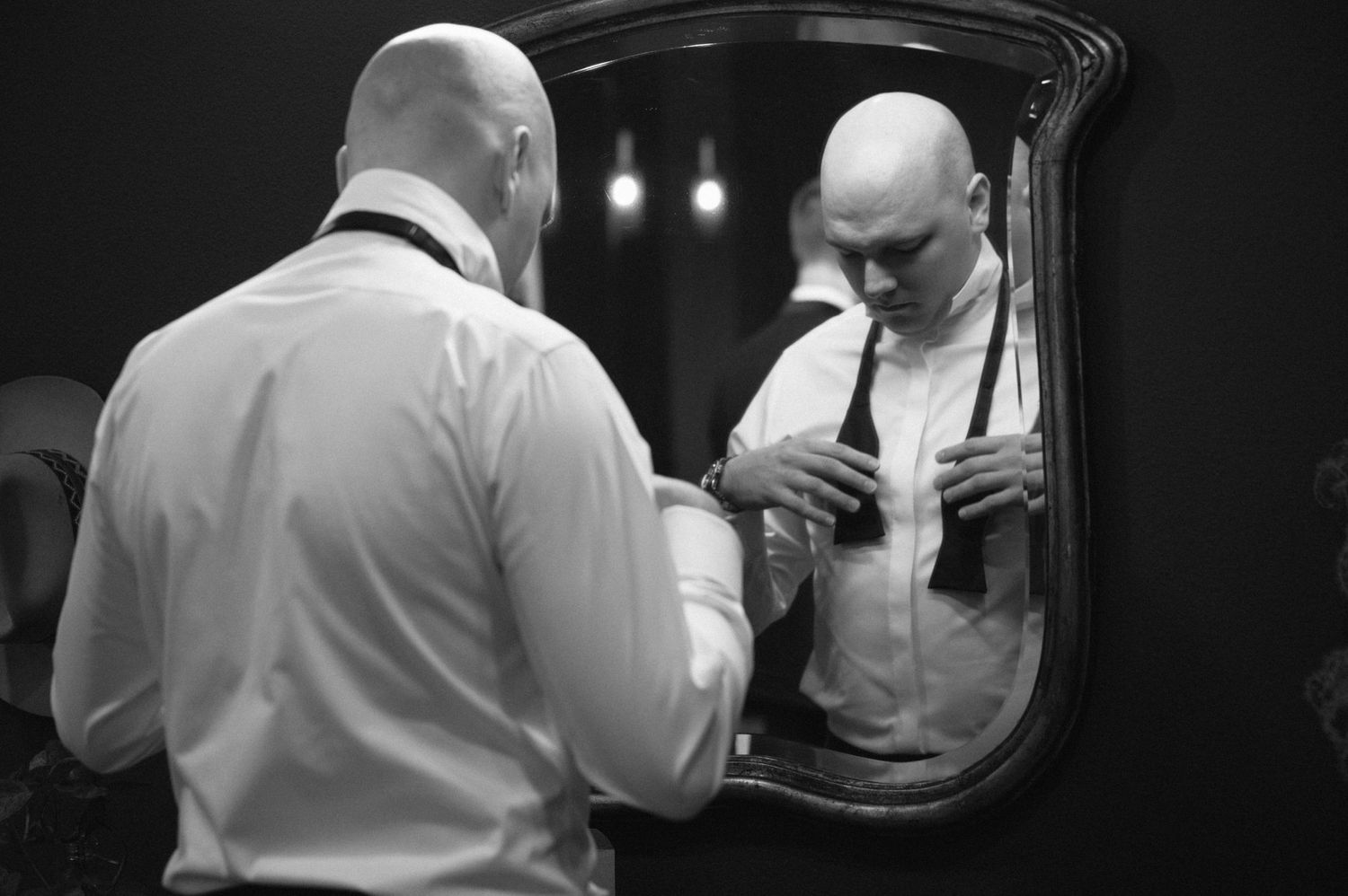 Black and white photograph showing a reflection in a mirror of someone adjusting their tie and formal attire.