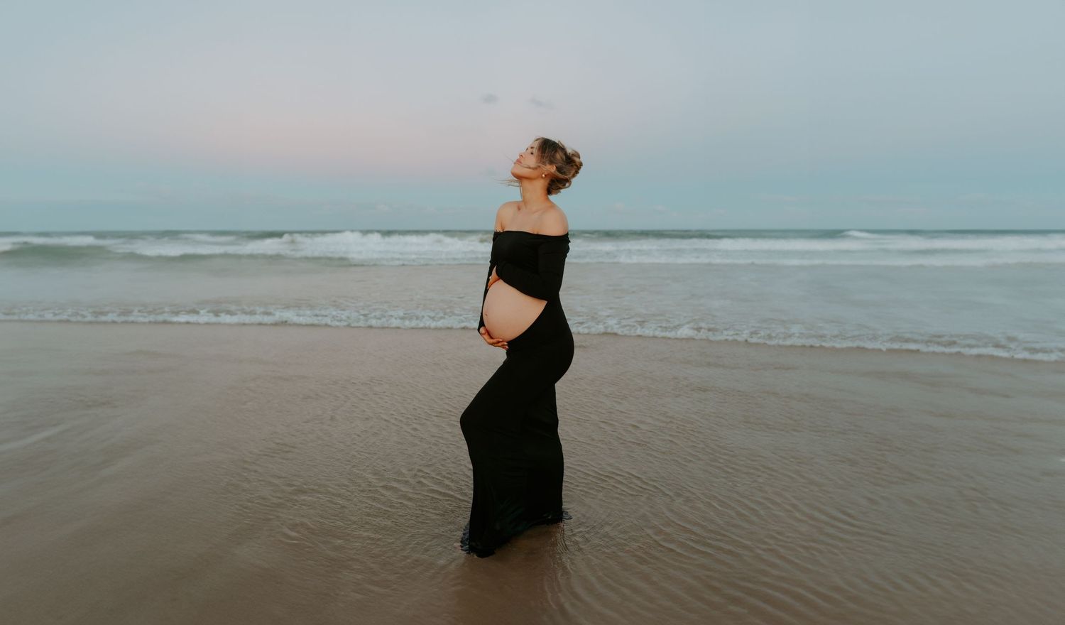 A silhouetted figure in black dress stands peacefully on a misty beach at dusk.