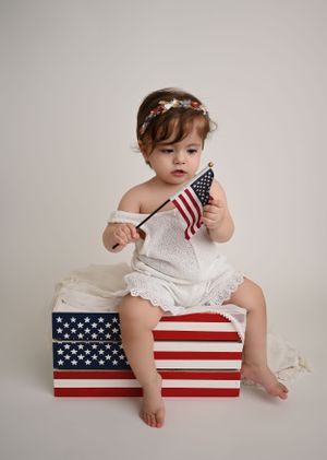 Patriotic-themed baby photoshoot in NYC, featuring a little girl holding an American flag with red, white, and blue props.