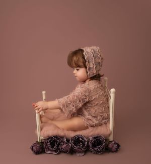 Rustic milestone photoshoot in NYC, capturing a baby girl in a lace romper and bonnet sitting in a miniature bed prop with soft florals.