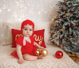 Adorable Christmas-themed baby portrait in NYC, with cozy red outfit, festive pillows, and a sparkling holiday tree backdrop.