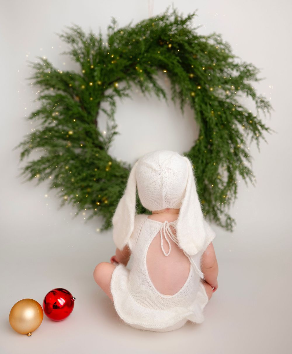 Festive holiday baby photoshoot in Brooklyn, NY, featuring a little one in white surrounded by a green wreath and Christmas ornaments.