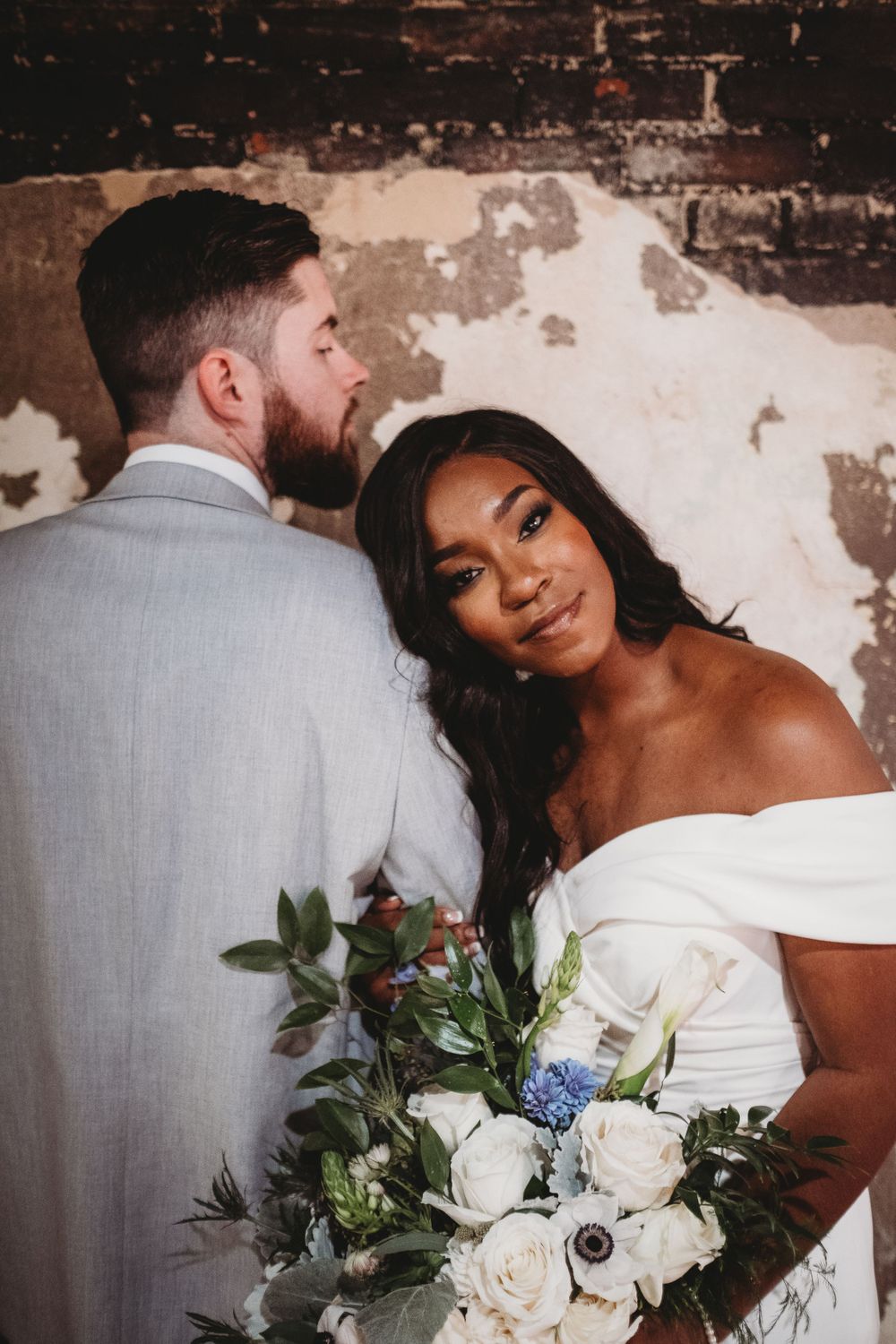 Elegant bride in an off-shoulder white gown holding white roses bouquet with groom in gray suit.