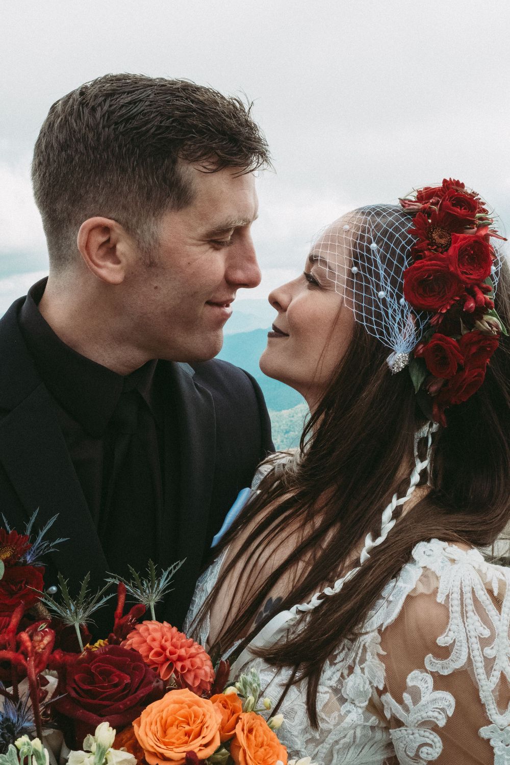 Bride wearing red floral crown and groom sharing an intimate moment with colorful bridal bouquet.