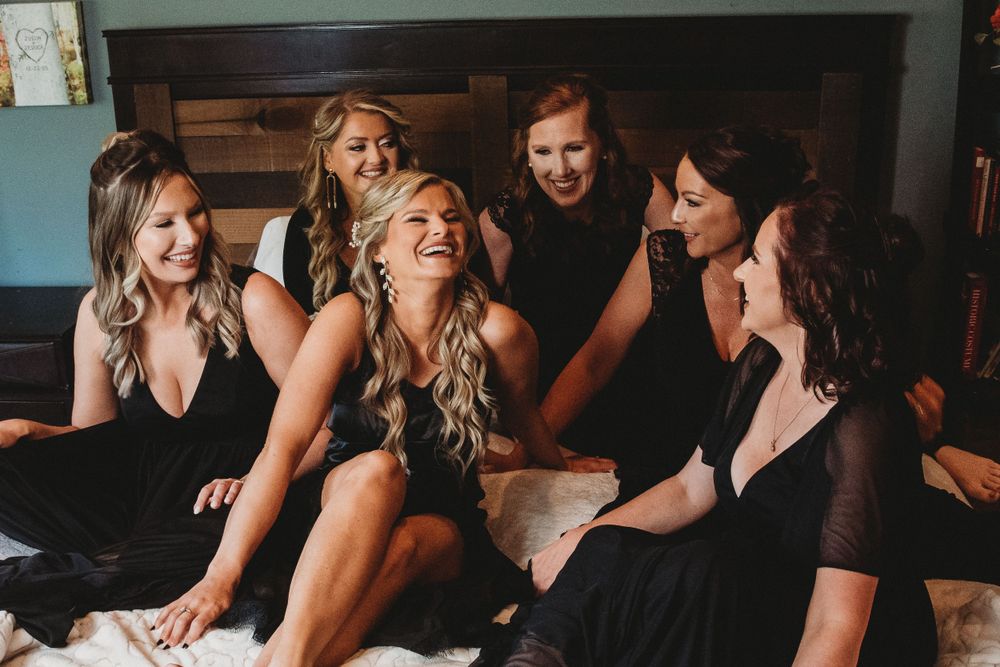 Six bridesmaids in black robes laugh together while sitting on bed during wedding preparations.